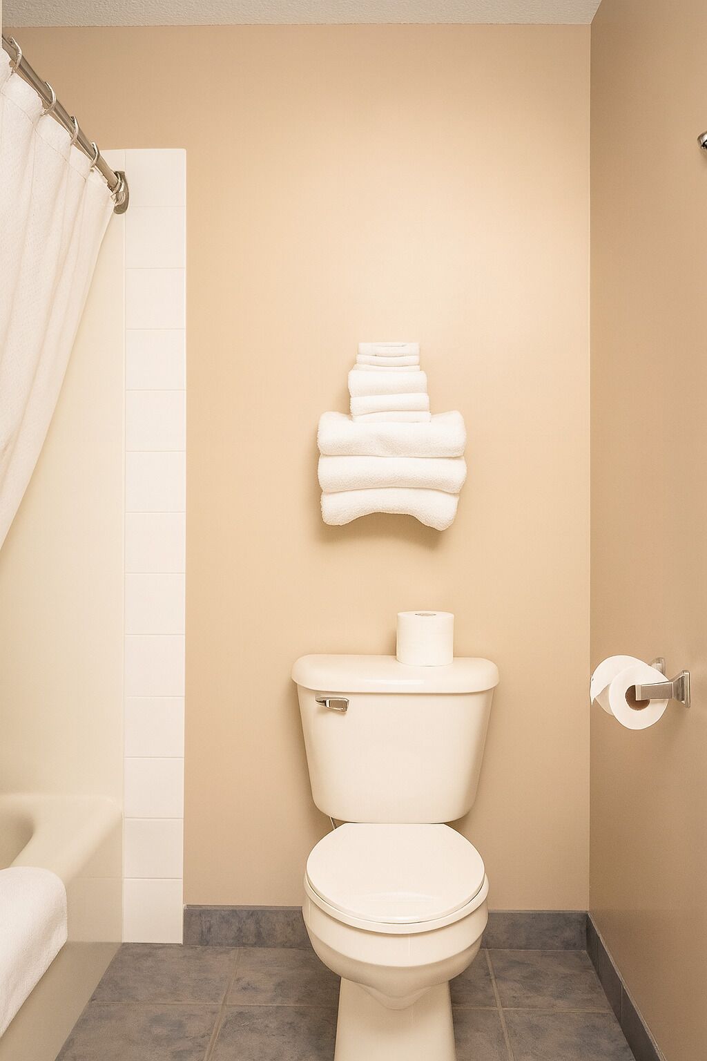 A bathroom with a toilet, a towel rack with folded towels, a shower curtain, and a toilet paper holder.