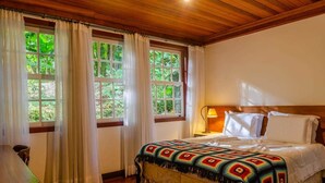 Une pièce chaleureuse avec un plafond en bois, un lit recouvert d’une couverture colorée, des rideaux blancs et une vue sur la verdure par les fenêtres.
