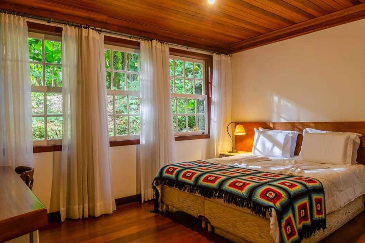 A cozy room with a wooden ceiling, a bed with a colorful blanket, white curtains, and a view of greenery through the windows.