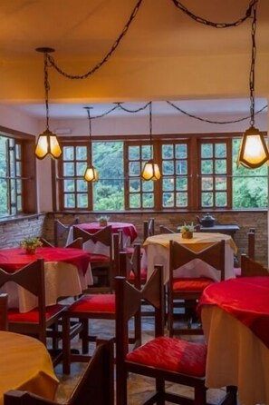 Ein Restaurant mit mehreren Tischen und Stühlen, großen Fenstern mit Blick auf Grünflächen und hängenden Pendelleuchten.