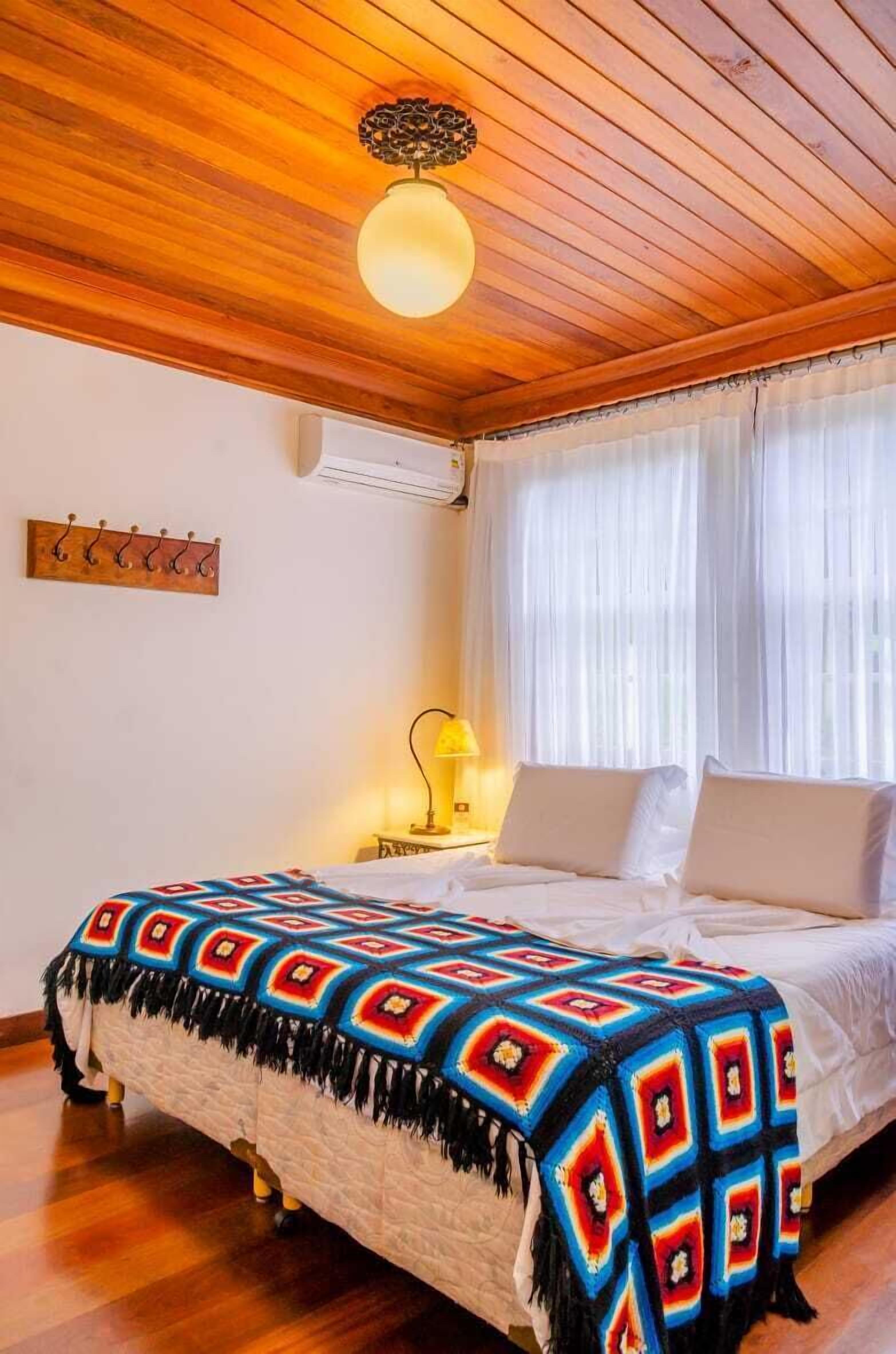 Une chambre confortable avec un plafond en bois, un lit recouvert d’une couverture colorée, une lampe de chevet et une fenêtre ornée de rideaux légers.
