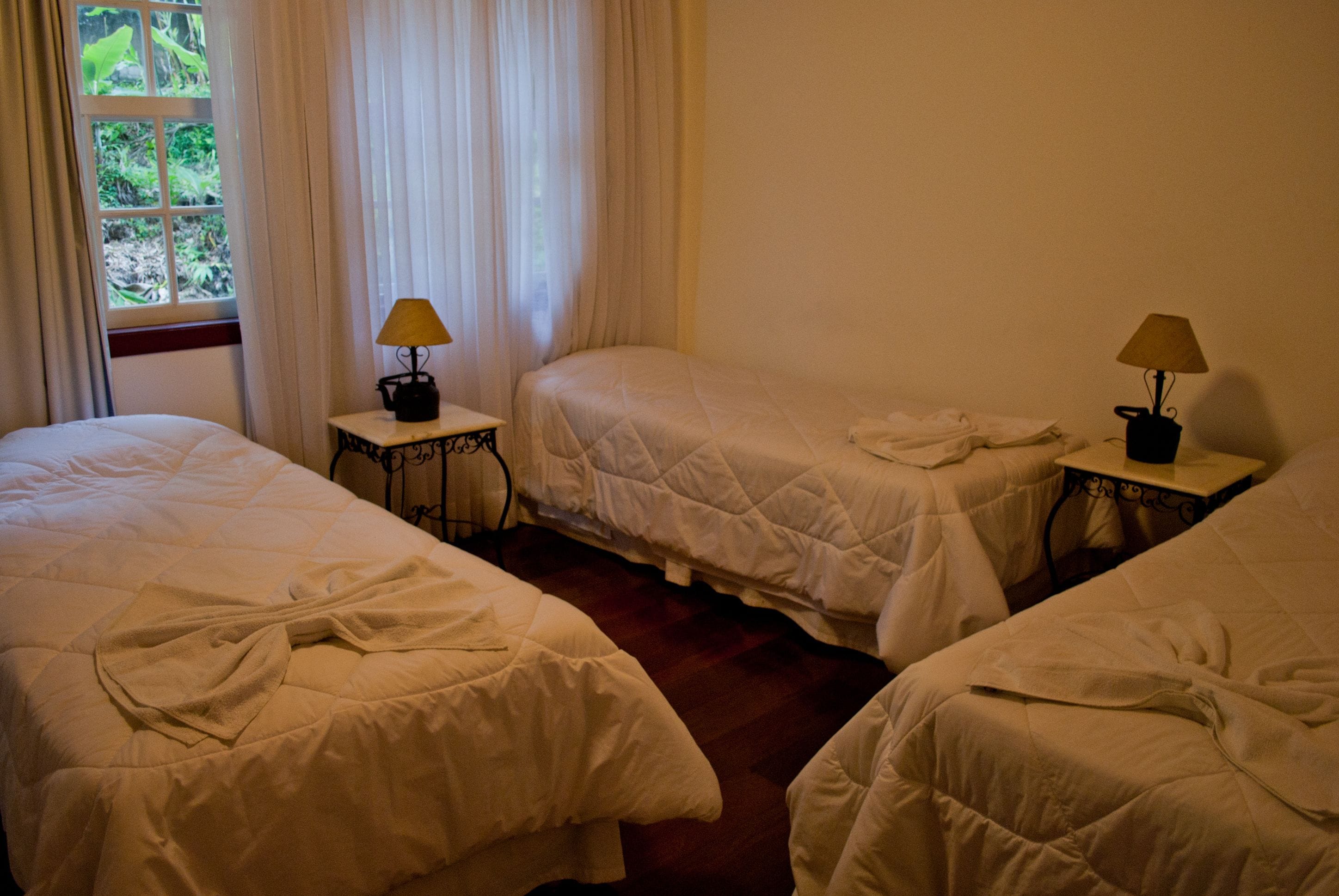Quarto de hotel com três camas de solteiro, cada uma com mesa de cabeceira e luminária.