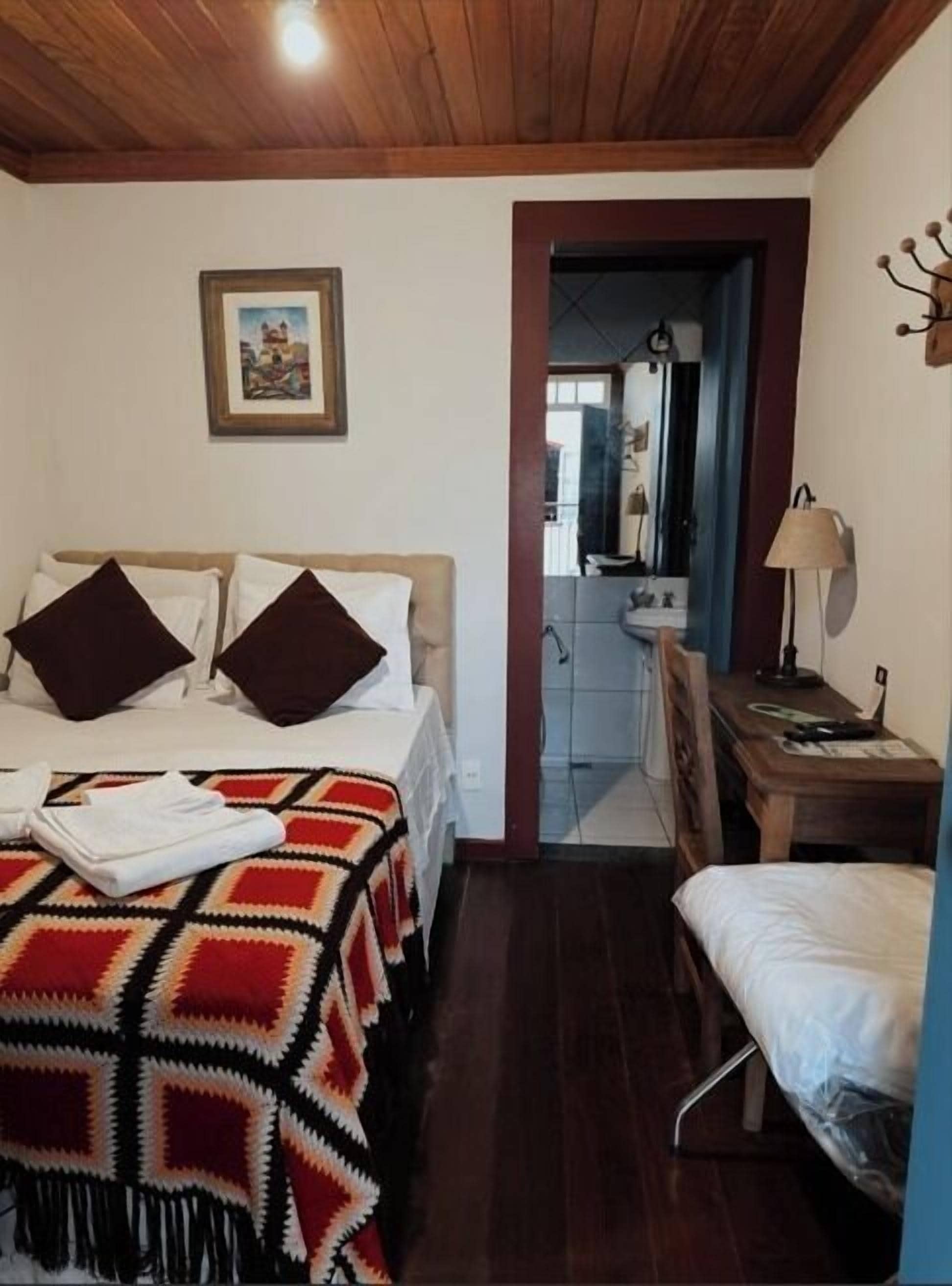 Quarto de hotel com cama, escrivaninha, cadeira, espelho e porta de acesso a um banheiro.