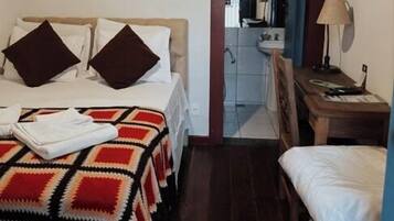 Quarto de hotel com cama, escrivaninha, cadeira, espelho e porta de acesso a um banheiro.