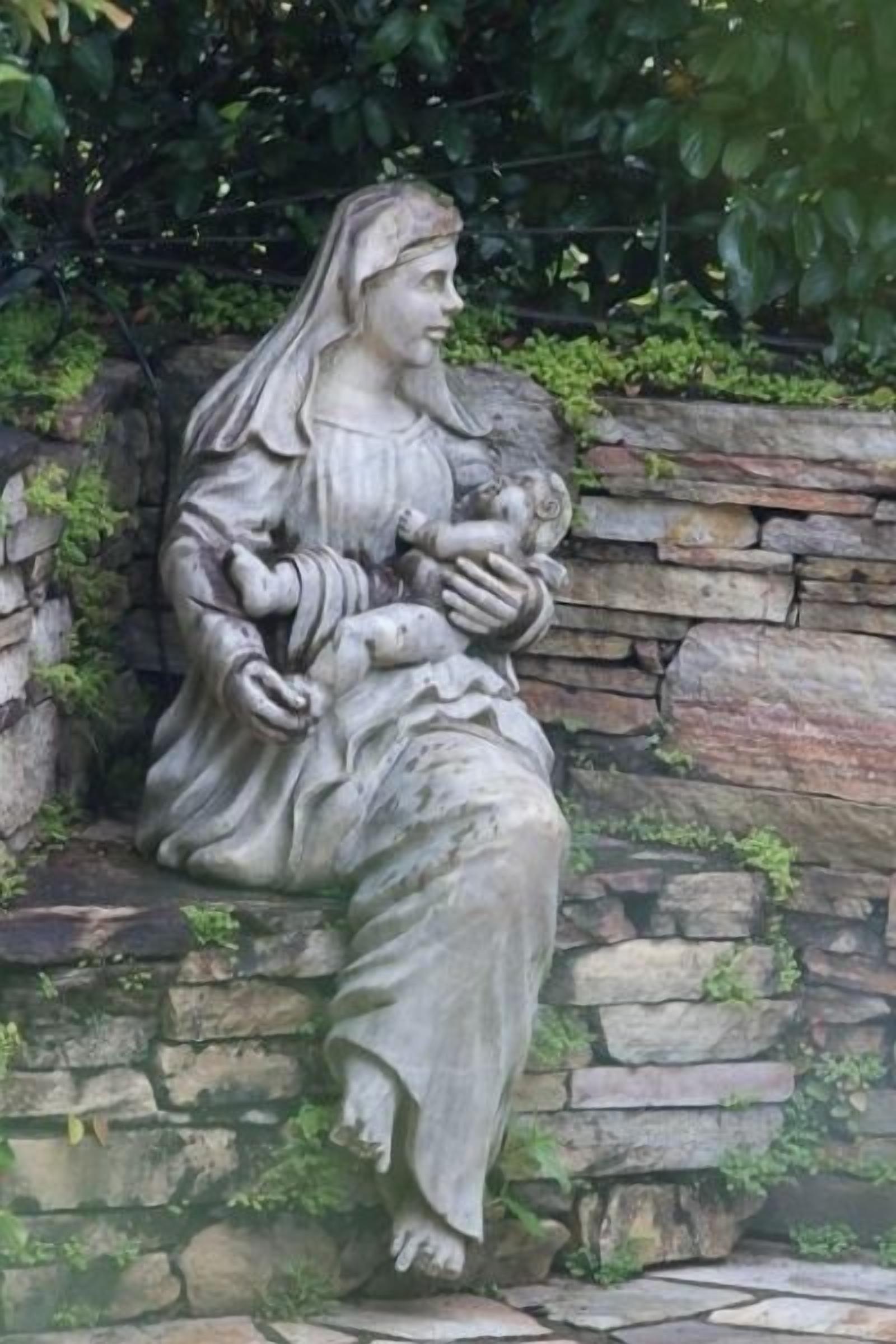 A stone statue of a seated figure holding a child, set against a stone wall with greenery in the background.
