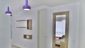 Quarto de hotel moderno com um espelho grande, cama com roupa de cama vermelha e área de jantar com mesa e cadeiras brancas.