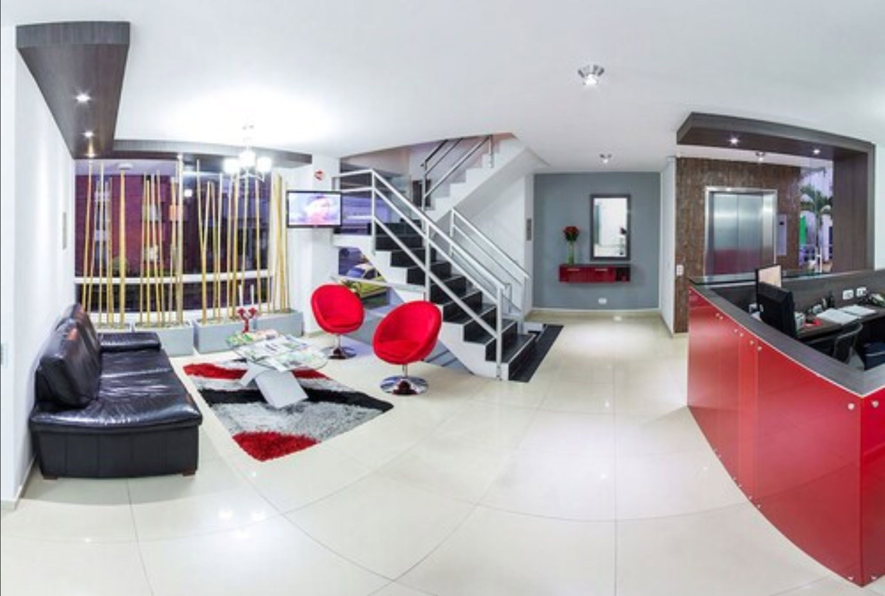 Sala de estar moderna com sofá de couro preto, cadeiras vermelhas, escada e balcão de atendimento vermelho.