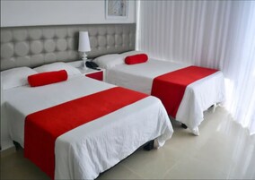 A hotel room with two beds, each with a white bedspread and red pillows. There is a nightstand with a lamp and a telephone between the beds. The room has a white headboard and a large window with sheer curtains.
