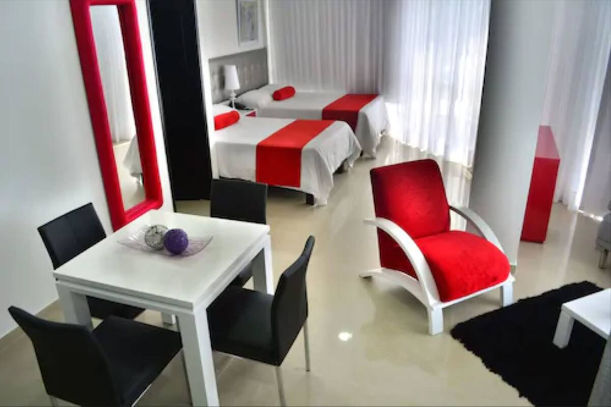 Ein Hotelzimmer mit zwei Betten, einem Esstisch mit Stühlen, einem roten Sessel und einem großen Spiegel.