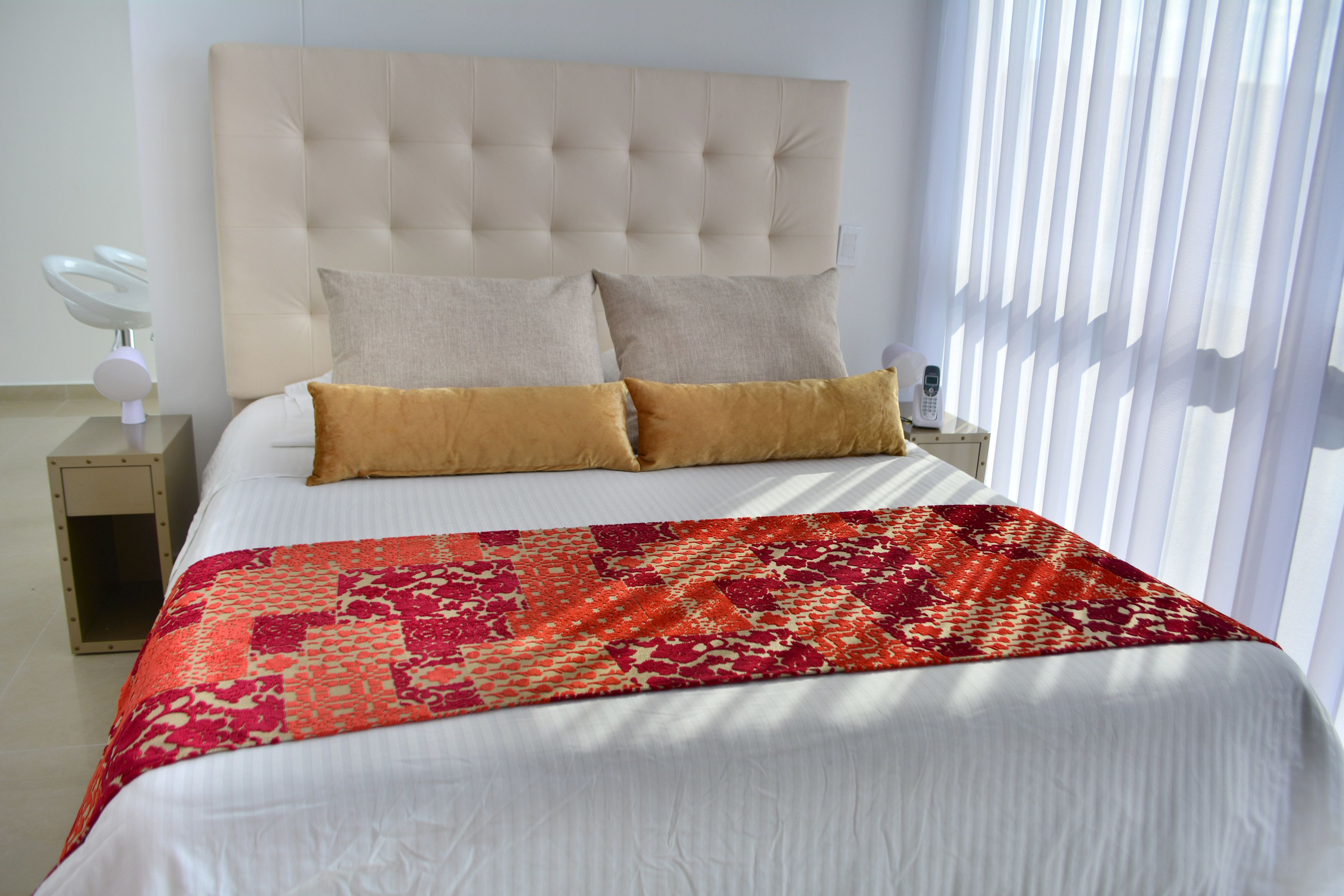 Een net opgemaakt bed met een geborduurde deken, een gestoffeerd hoofdbord en twee nachtkastjes met een lamp en een telefoon.