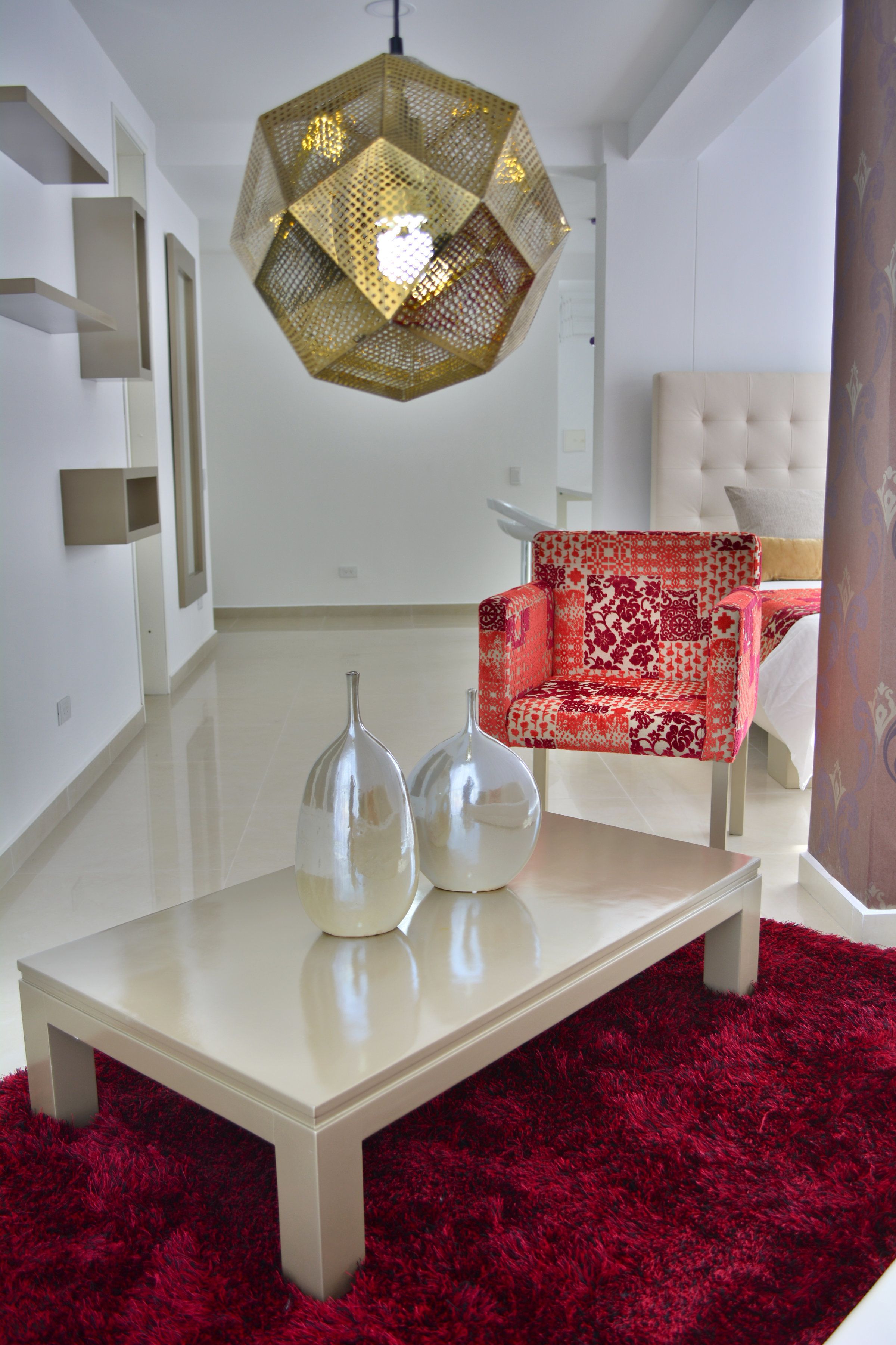 Ein modernes Wohnzimmer mit einem rot gemusterten Ohrensessel, einem weißen, gepolsterten Sofa, einer geometrischen Pendelleuchte und einem roten, hochflorigen Teppich.