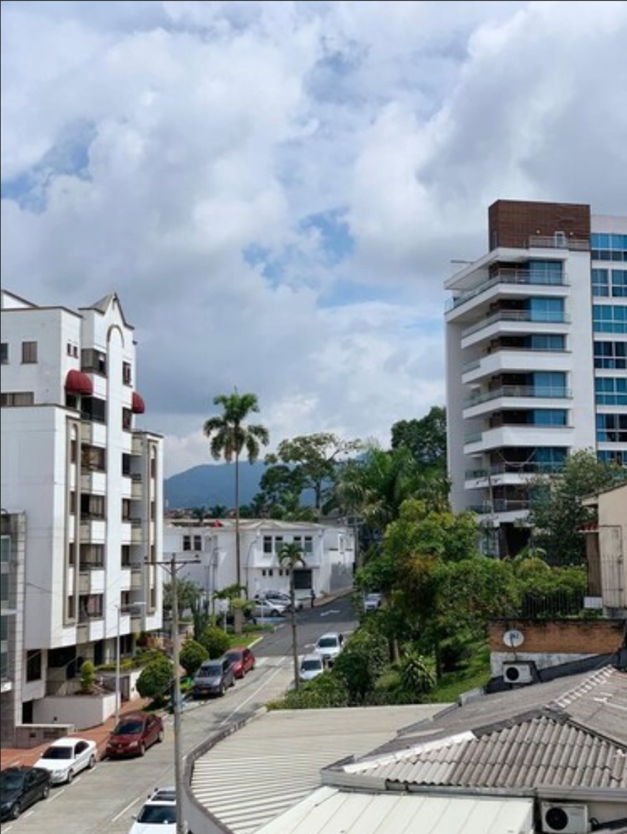 Un edificio residencial de varios pisos con balcones, una calle con coches aparcados y una montaña al fondo.