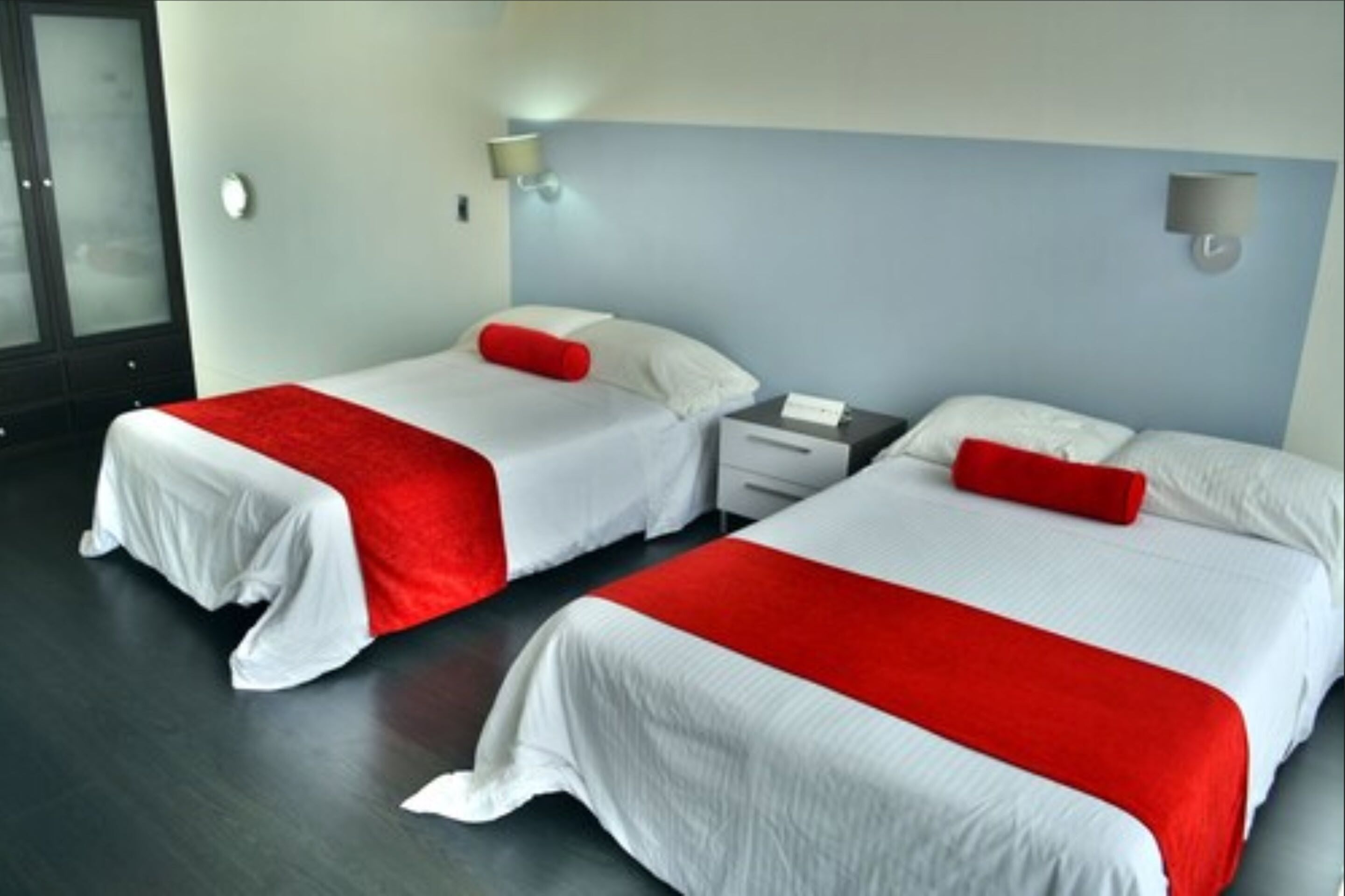 A hotel room with two beds, each with white bedding and red accents, a bedside table with a lamp, and a nightstand with a phone.