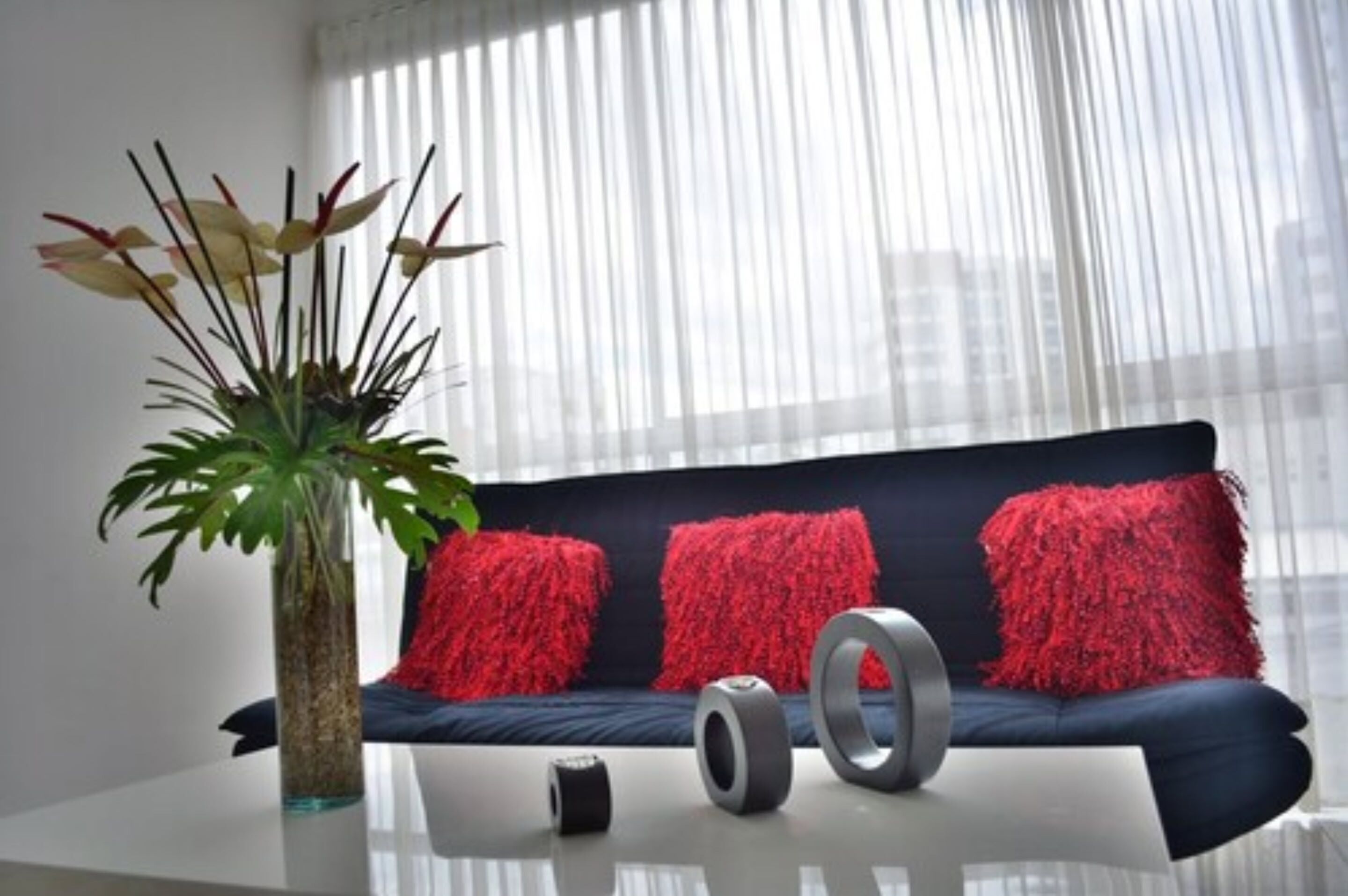 Un salón moderno con un sofá azul oscuro, cojines rojos, un florero de vidrio con una planta y dos objetos cilíndricos sobre una mesa de vidrio.