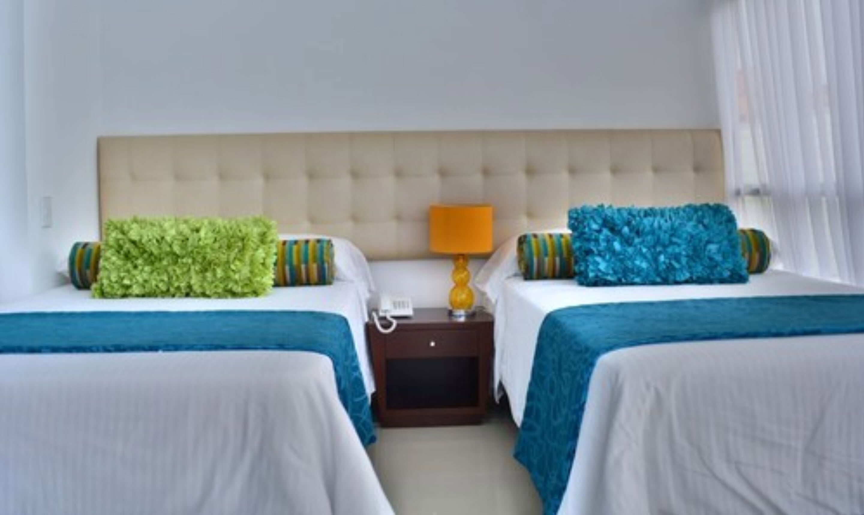 A hotel room with two beds, each with a blue bedspread and a green and striped pillow. There is a nightstand with a lamp and a phone between the beds.