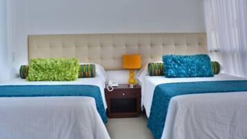 A hotel room with two beds, each with a blue bedspread and a green and striped pillow. There is a nightstand with a lamp and a phone between the beds.