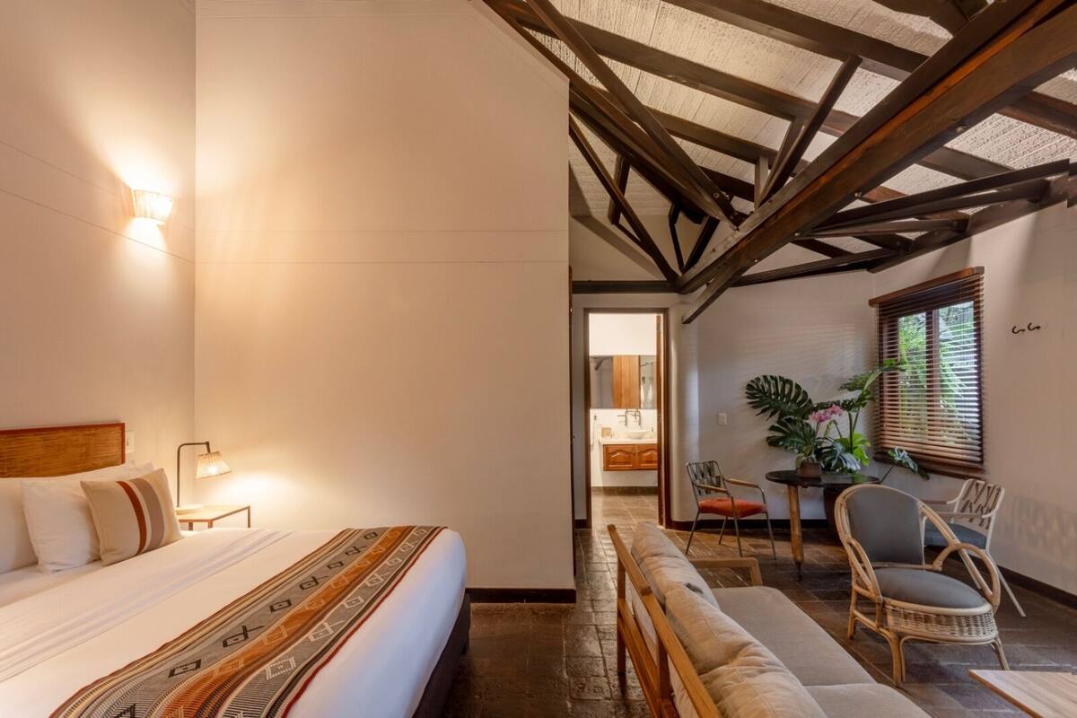 A hotel room with a bed, a nightstand, a lamp, a sofa, a dining table with chairs, and a view of the bathroom.