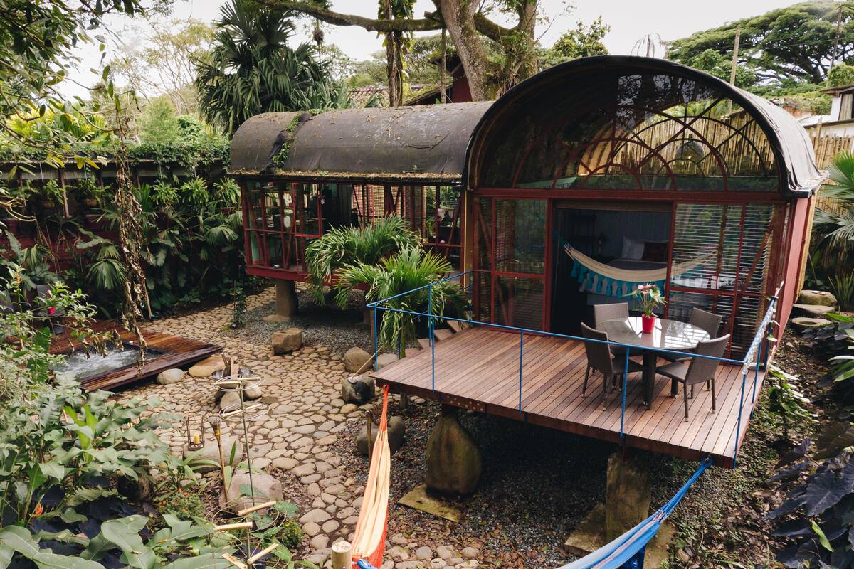 A wooden deck with a glass enclosure, surrounded by lush greenery and a stone pathway.