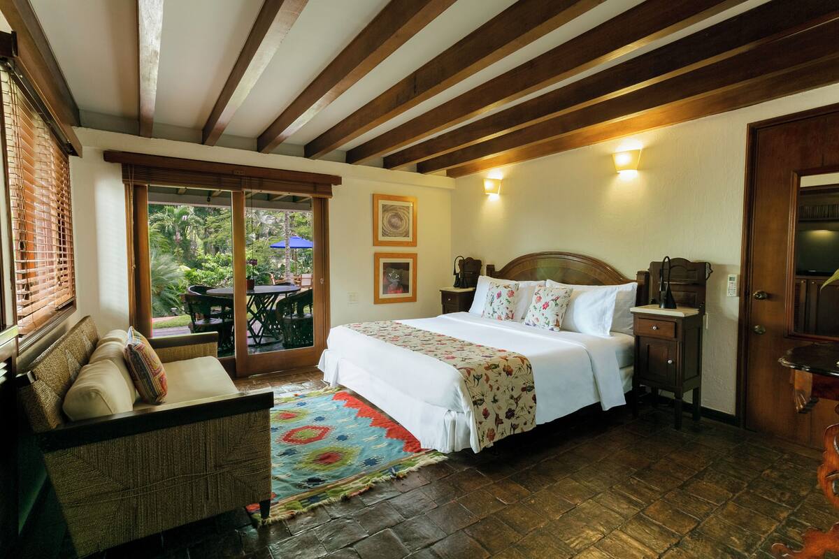 A bedroom with a large bed, wooden ceiling beams, a small table, and a view of the outdoors.