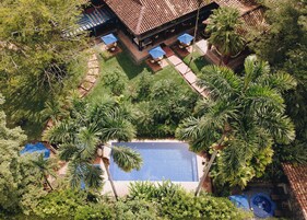 An aerial view of a resort with a swimming pool surrounded by palm trees, outdoor seating, and a covered area with blue umbrellas.