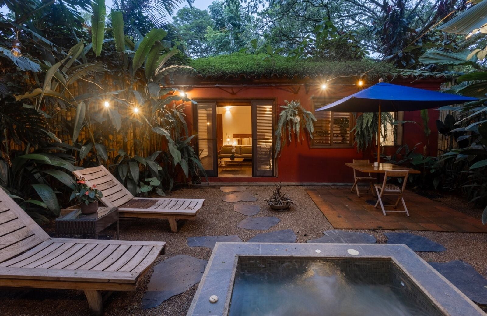 A cozy outdoor patio with wooden lounge chairs, a small pool, and a dining area with a blue umbrella.