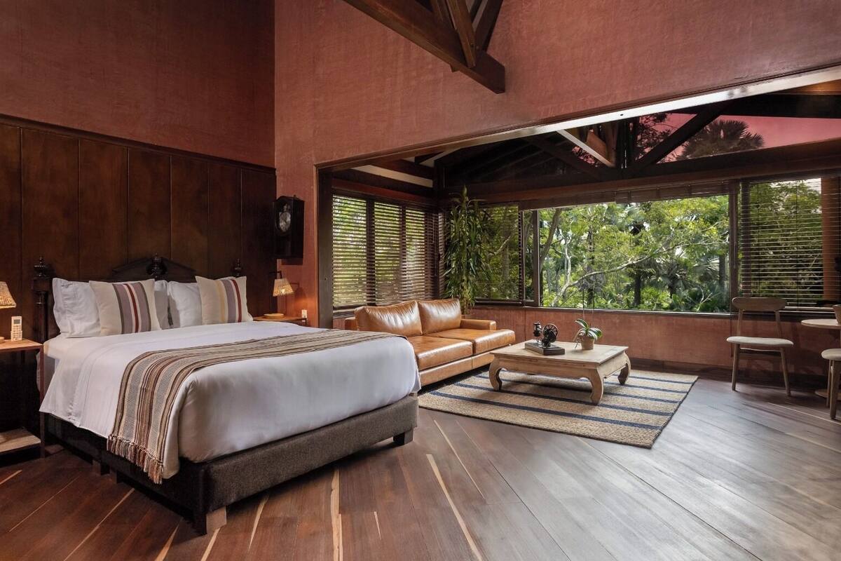 A spacious bedroom with a large bed, a leather sofa, a coffee table, and a view of lush greenery through large windows.