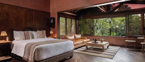 Un dormitorio amplio con una cama grande, un sofá de cuero, una mesa de centro y vistas a vegetación exuberante a través de ventanas grandes.