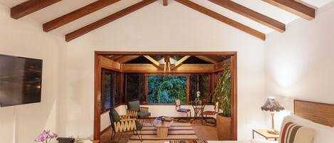 A spacious room with a high ceiling, wooden beams, a large bed, a seating area with chairs, a small table, and a view of greenery through the windows.