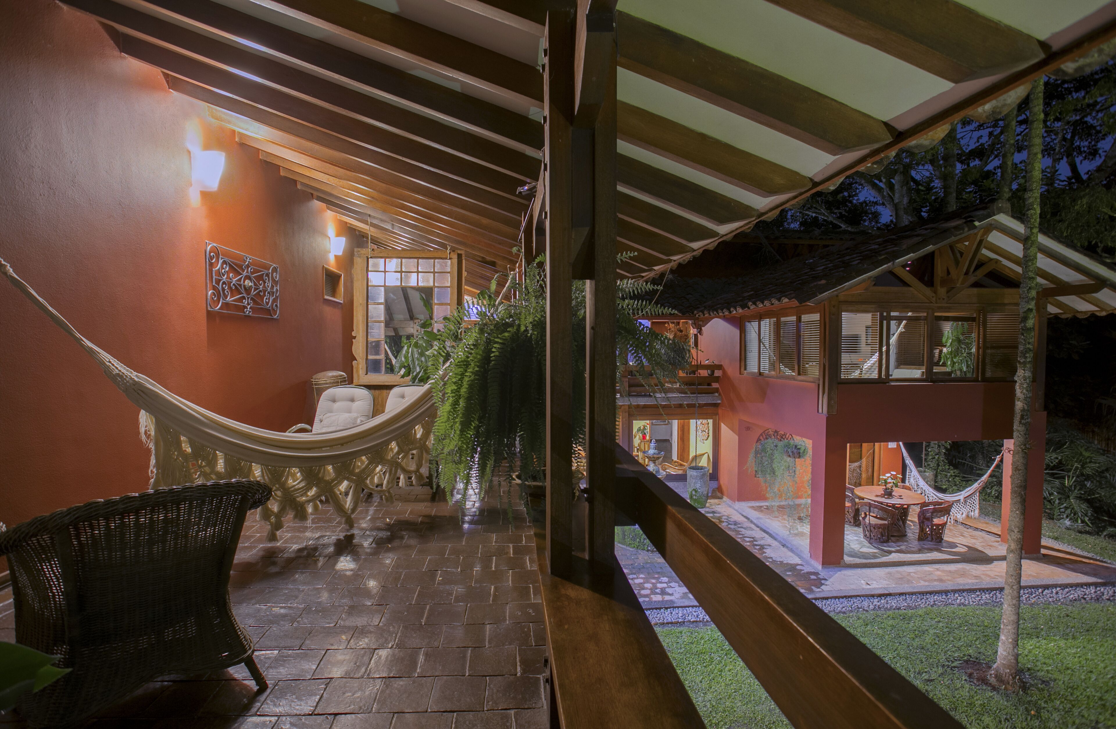 Un espacio al aire libre techado con una hamaca, muebles de mimbre y vista a un edificio de estructura de madera y techo de paja.