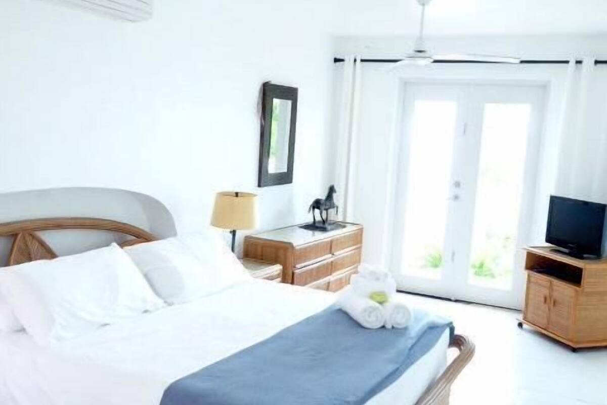 Un lit bien fait, recouvert d’une couverture bleue, une table de chevet en bois avec une lampe, un tableau encadré et un téléviseur posé sur une commode en bois.