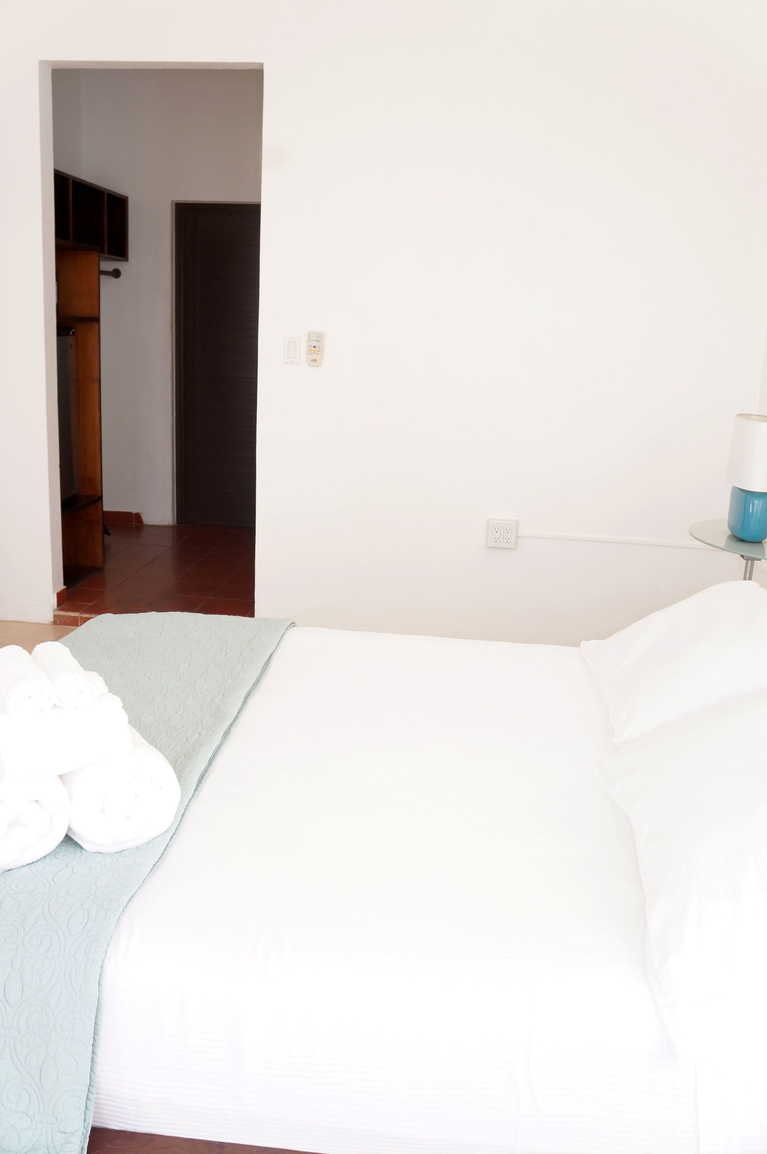 A bedroom with a bed, a bedside table with a lamp, and a door leading to another room.