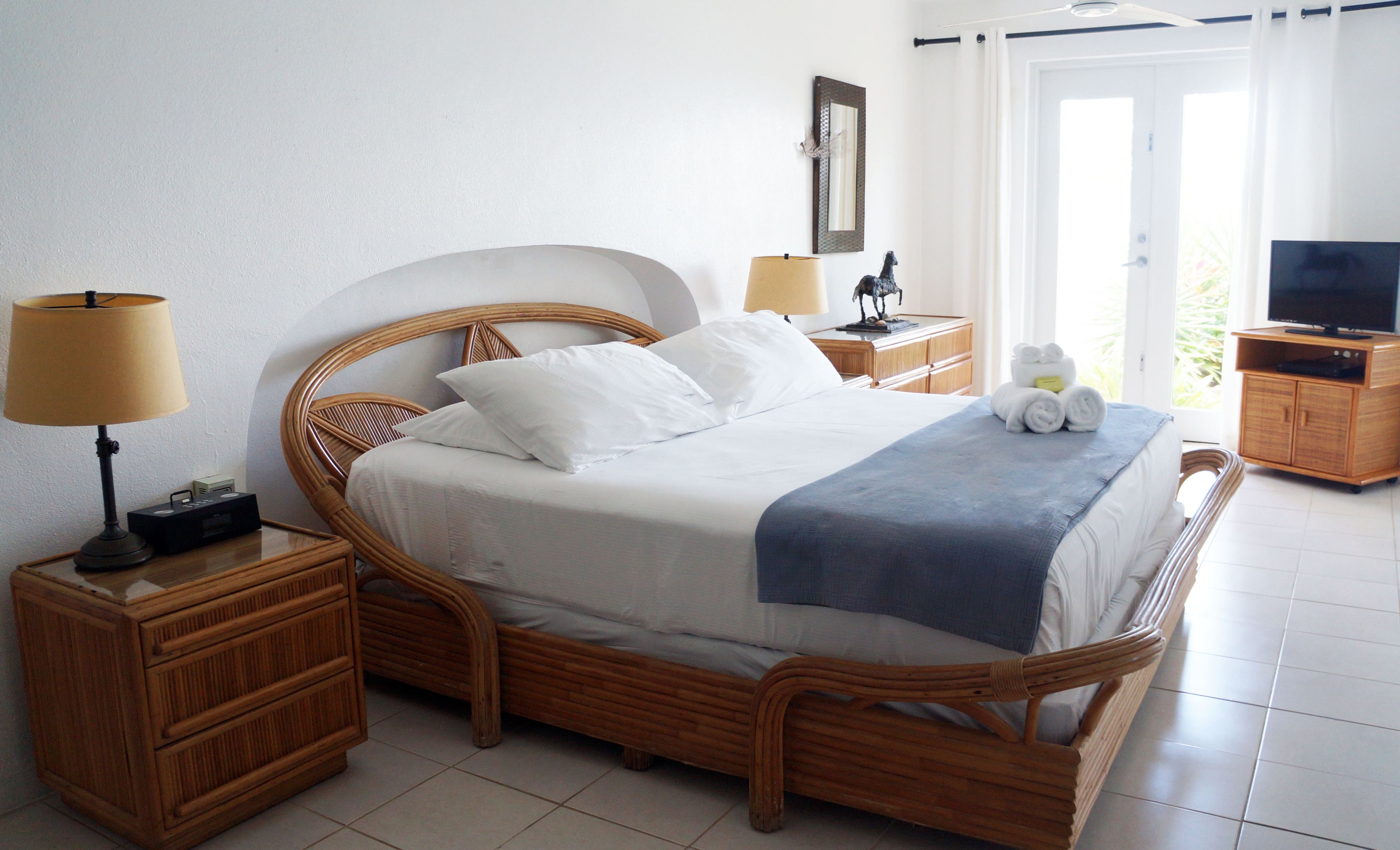 A neatly made bed with white linens, two bedside tables with lamps, a wooden nightstand, a television on a wooden cabinet, and a large window with curtains.