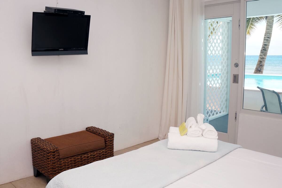 Une chambre d’hôtel dotée d’un téléviseur à écran plat fixé au mur, d’un banc en osier avec un coussin marron, d’un lit avec du linge de maison blanc et d’une vue sur l’océan par une porte vitrée.