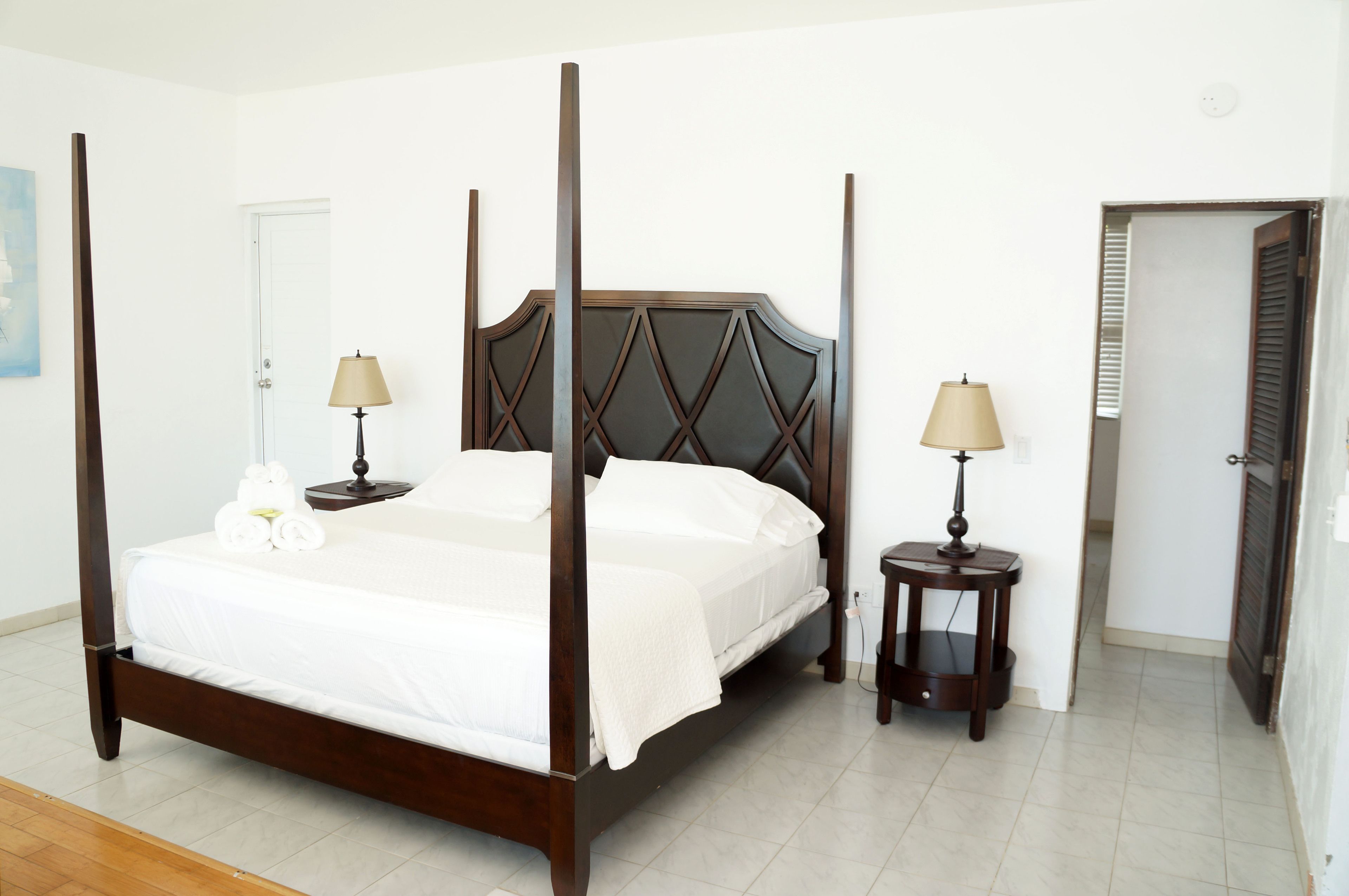 Una cama con dosel, ropa de cama blanca, dos mesitas de noche con lámparas y una puerta que da a otra habitación.
