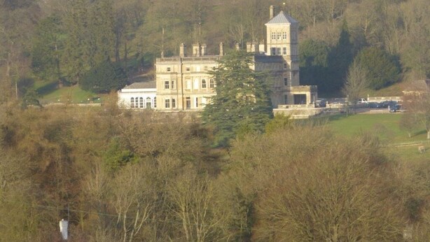 Un edificio grande e histórico con varios pisos y una torre destacada, rodeado de árboles densos y un paisaje montañoso.
