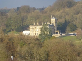 A large, historic building with multiple stories and a prominent tower, surrounded by dense trees and a hilly landscape.