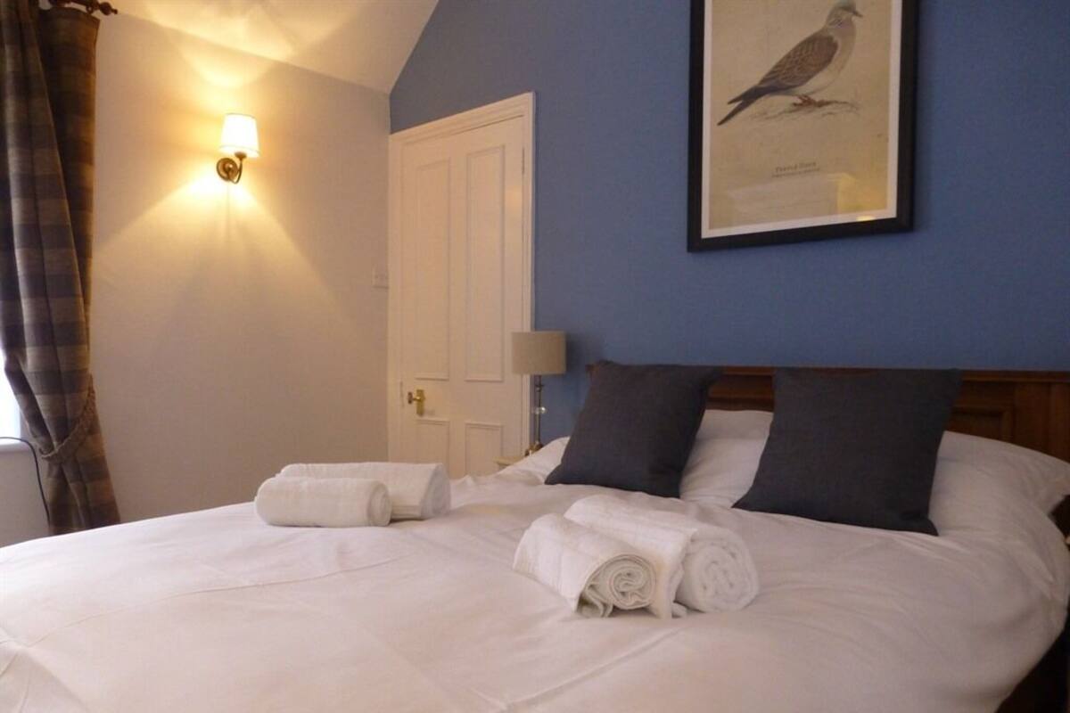 A neatly made bed with white linens and rolled towels, a framed bird illustration on the wall, and a door in the background.