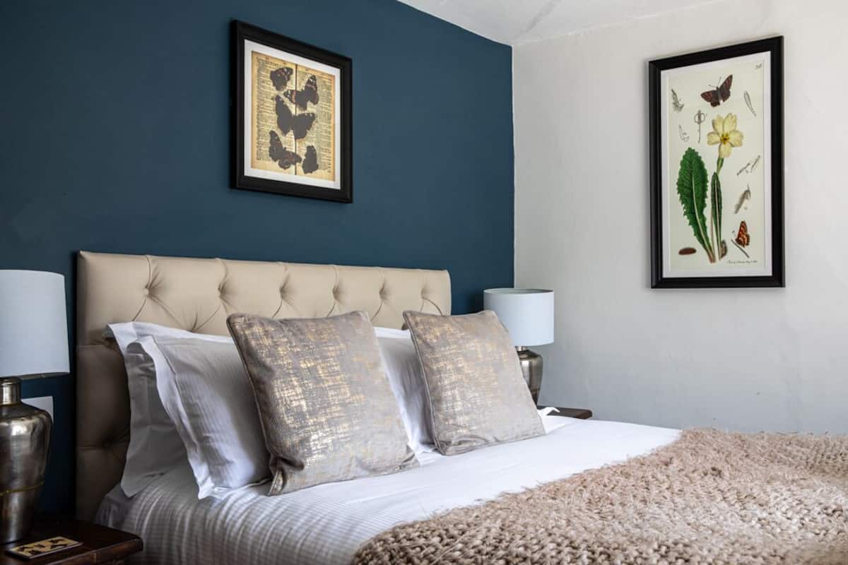 A bedroom with a tufted headboard, a large bed with white linens, two decorative pillows, a beige throw blanket, and two wall-mounted lamps.
