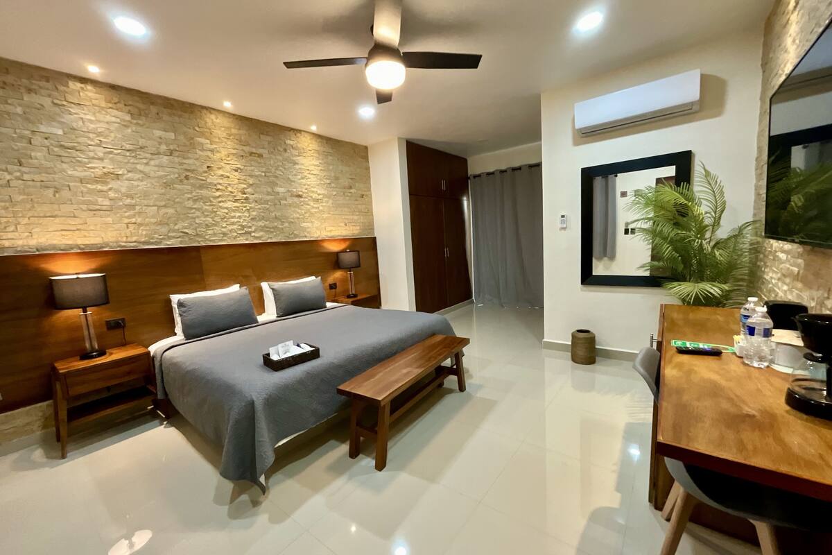 Habitación de hotel moderna con pared de piedra decorativa, cabecero de madera, cama con ropa de cama gris, mesita de noche con lámpara, banco de madera, escritorio con silla, planta en maceta y ventilador de techo.