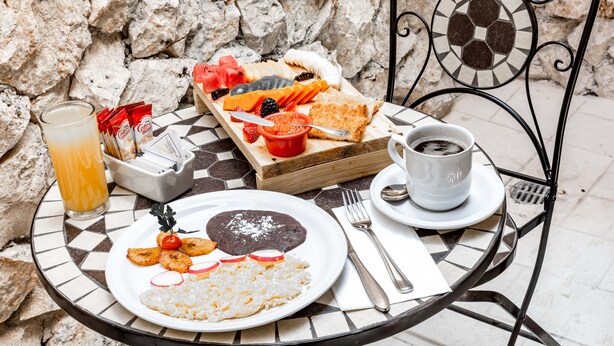 Une table ronde est dressée pour le repas, avec un mur de pierres en arrière-plan. On y trouve une assiette de riz, de haricots et de légumes, un plateau avec divers fruits et une tasse de café.