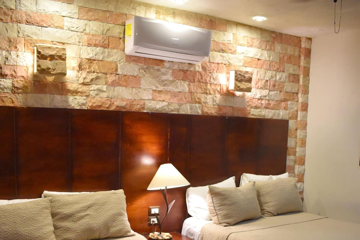 Habitación de hotel con dos camas, cabecero de madera, mesita de noche con lámpara y un equipo de aire acondicionado instalado en la pared.