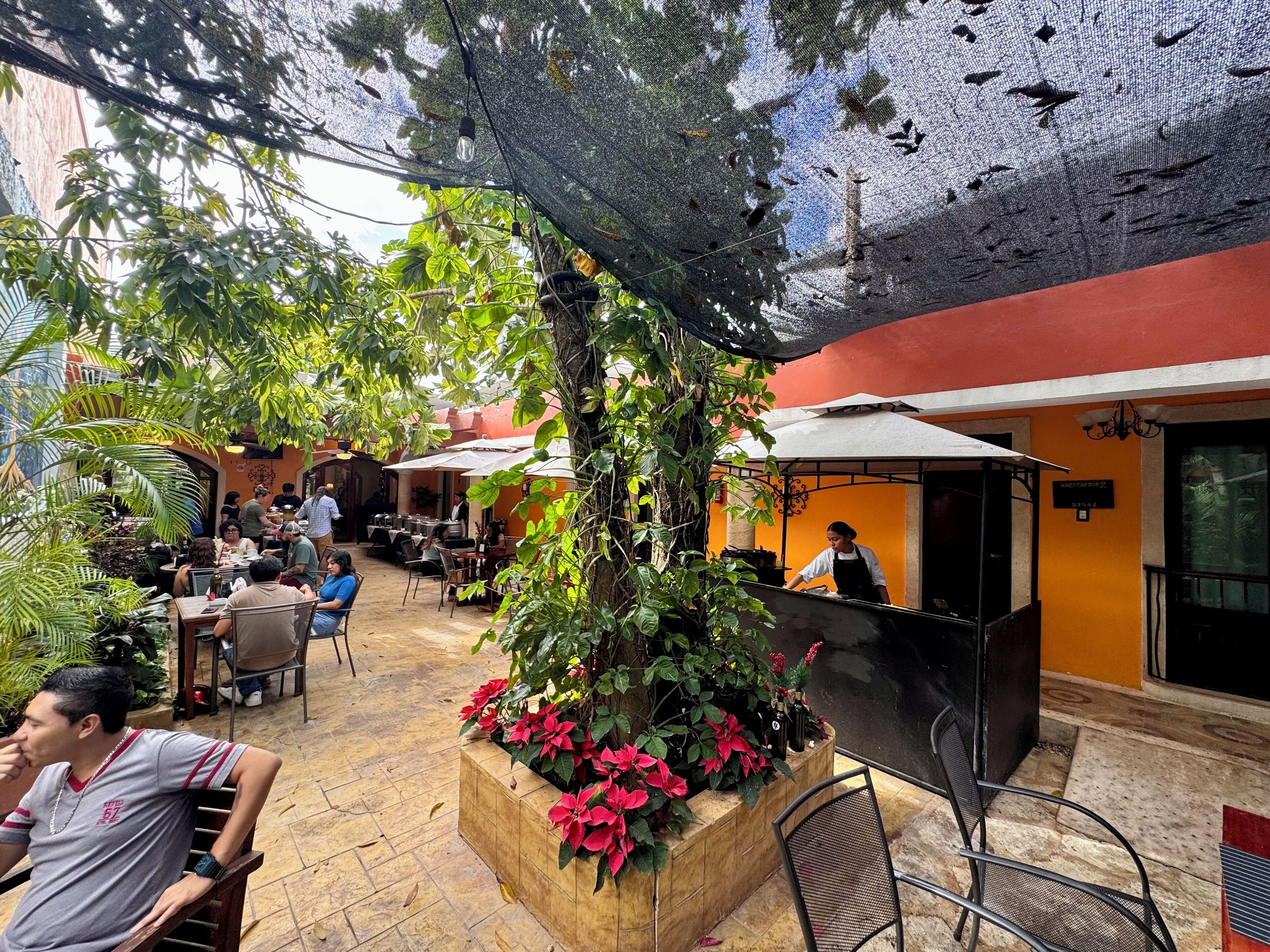 An outdoor cafe area with tables and chairs, surrounded by greenery and a decorative planter with flowers.