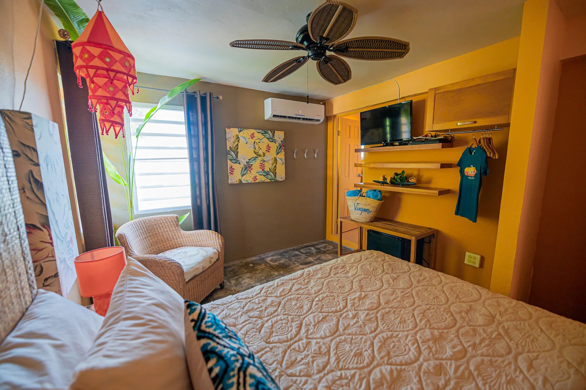 A hotel room with a bed, a chair, a ceiling fan, an air conditioning unit, a television, and a wall-mounted shelf with a towel and a bag.