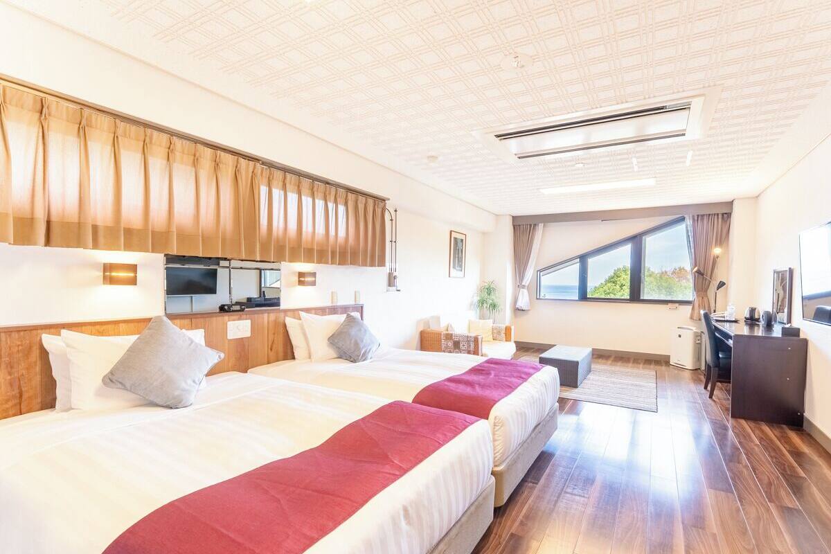 A hotel room with two beds, a desk, a chair, a small table, a TV, and a window with a view.