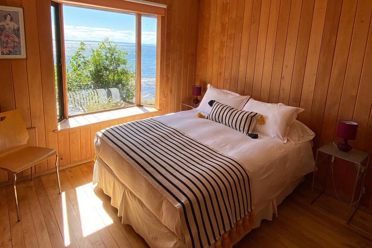 Un dormitorio con paneles de madera, una cama grande, una silla, una mesita de noche y una ventana con vistas al mar.