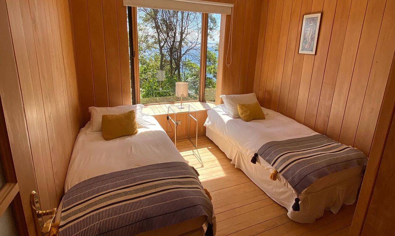 Una habitación con dos camas, paredes de madera, un ventanal con vistas a árboles y una mesita con lámpara.