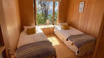 Una habitación con dos camas, paredes de madera, un ventanal con vistas a árboles y una mesita con lámpara.