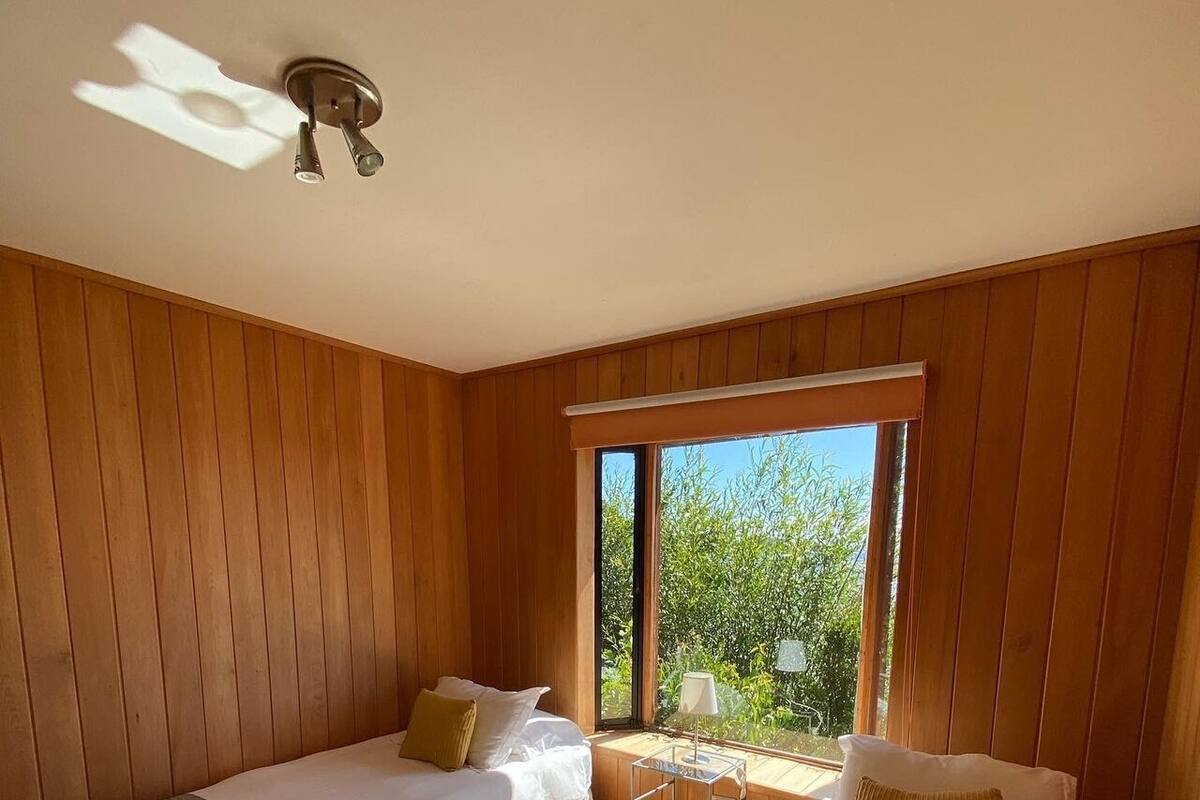 Habitación con dos camas, paredes de madera, una mesita pequeña y ventana con vista a la vegetación.