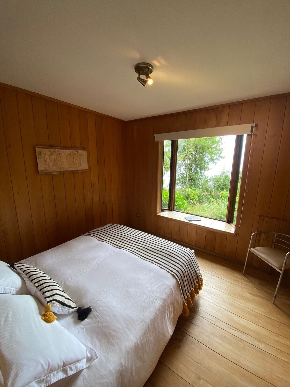 A bedroom with a wooden floor, a bed with striped bedding, a window with a view of greenery, and a wall-mounted light fixture.