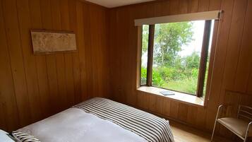 A bedroom with a wooden floor, a bed with striped bedding, a window with a view of greenery, and a wall-mounted light fixture.